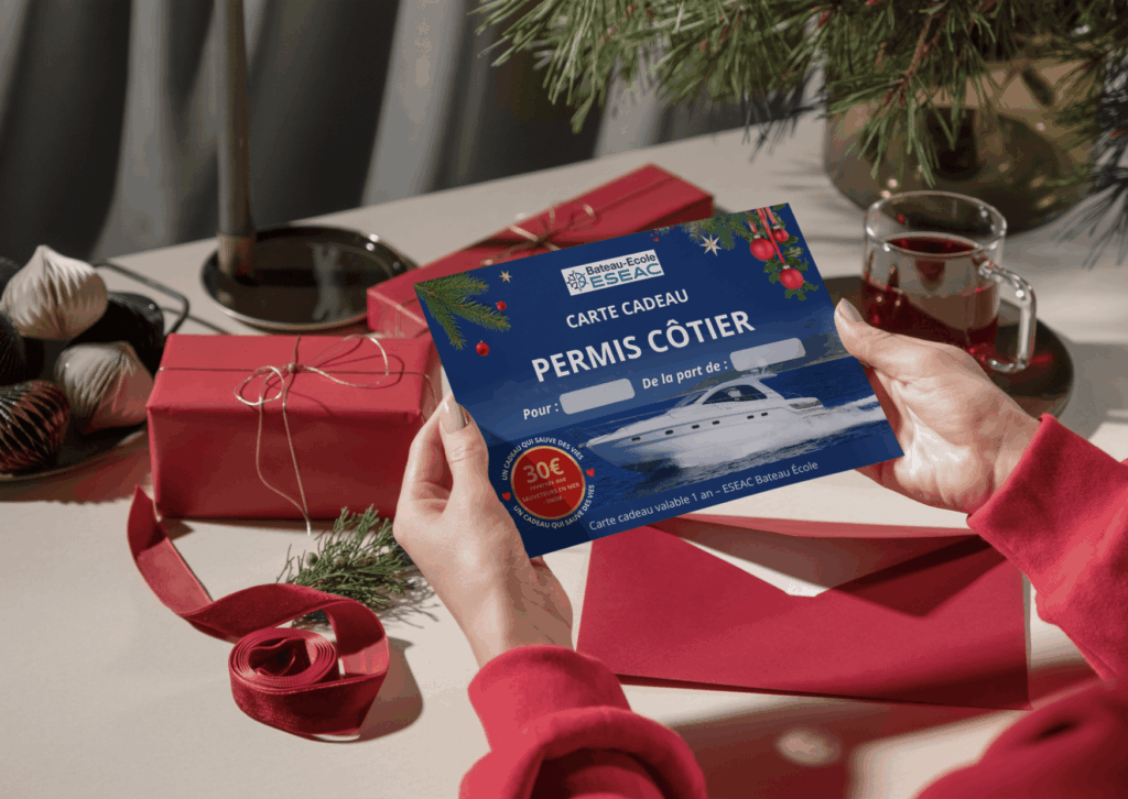 Carte cadeau de Noel 2025 ESEAC Bateau École pour le permis côtier, présentée dans une enveloppe rouge sur une table décorée de Noël. Idée cadeau originale pour apprendre à naviguer en mer avec un moniteur de 30 ans d'expérience et bénévole à la SNSM. Don de 30euros reversés aux sauveteurs en mer pour chaque carte cadeau achetée.